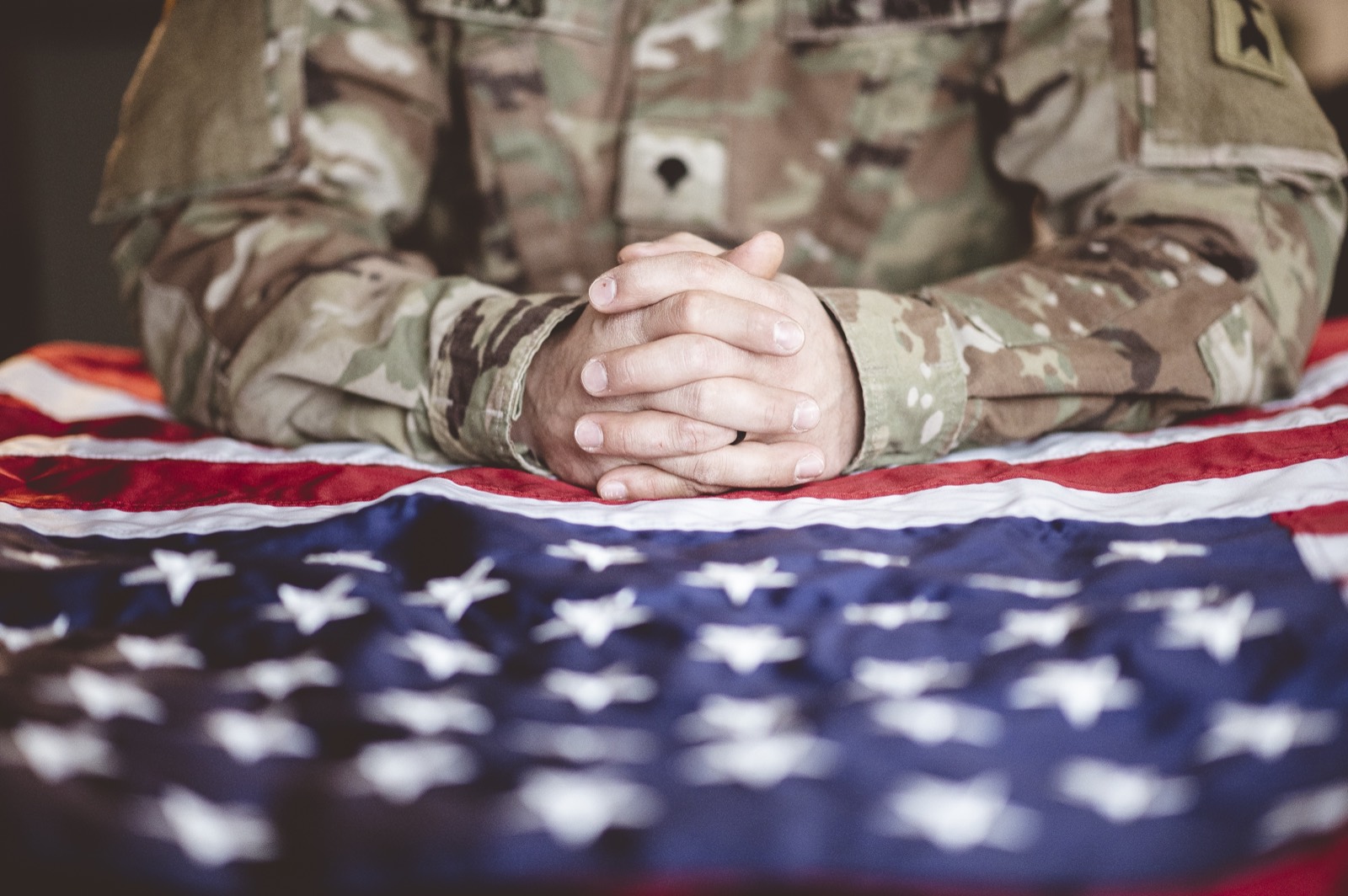 A soldier in uniform with hands clasped resting on an American flag, honoring those who served