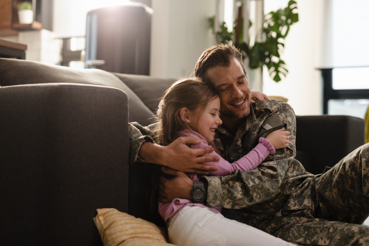 A smiling service member in uniform embracing his young daughter at home